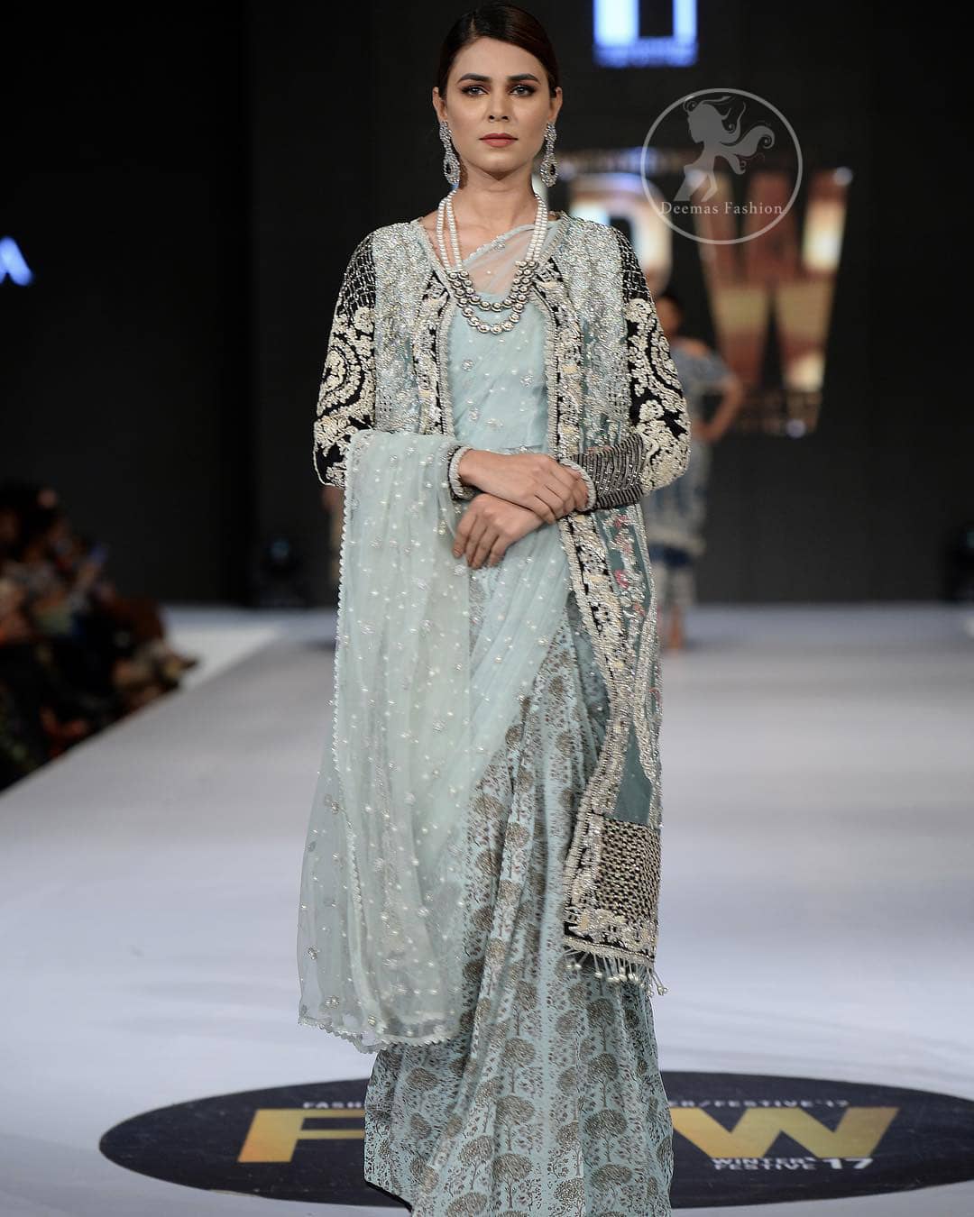 This outfit is a timeless beauty. The blouse is covered with gown embellished with silver gold kora dabka, Sequins and swarovski crystals.This exquisite gown is decorated with embroidered black applique and tassels on hemline.It comes with lehenga style bottom. This Outfit is beautifully coordinated with strip of material that wrapped around the blouse and draped across the front and over one shoulder?