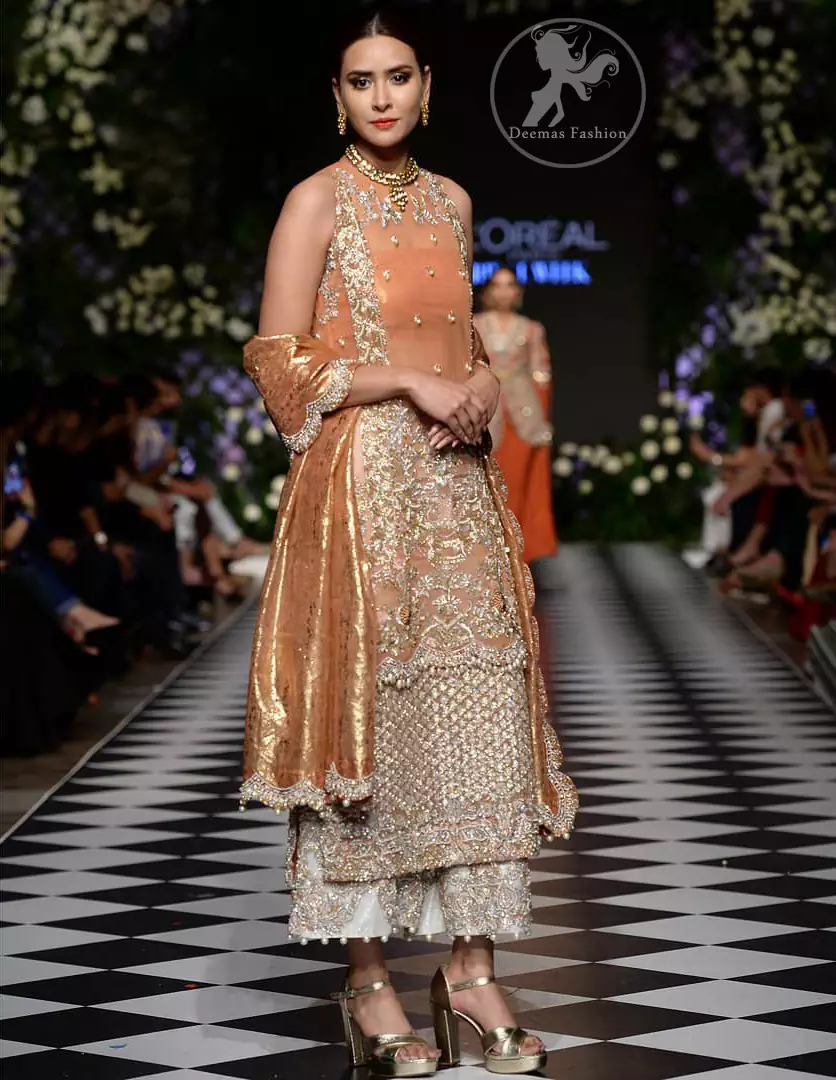 This outfit is stunningly perfect for any evening ensemble. It is paired with a sleeveless double layered shirt in organza fabric which is extensively hand worked in crystals and zardozi. This dress is beautifully sculptured with dull golden floral embroidery, adorned with cutwork borders embellished with tassels, kora, dabka, pearl and sequins work all over. It comes with embroidered trousers. It is coordinated with tissue dupatta which is furnished with four sided scalloped border.