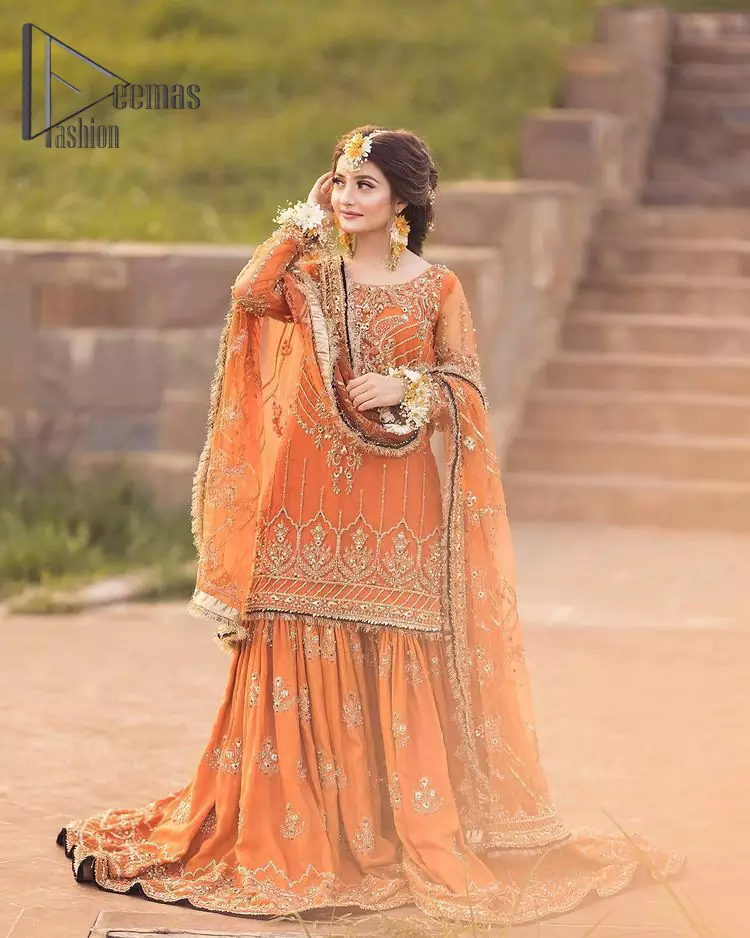 Time to treat yourself with an orange article of Deemas Fashion. This orange heartiest mealtime article will twirl your heart.  Prettify your Mehndi look with this orange short shirt that is embellished with tilla, kora and dabka work. Its rounded neckline with full sleeves gives you a genuine feeling of a bride. It is synchronized with flayer orange sharara that has an embellished border and spray of tiny floral motifs that gives a super aesthetic look to this dress. Finish this look with a net dupatta that has four sides embellished border and Kiran work.